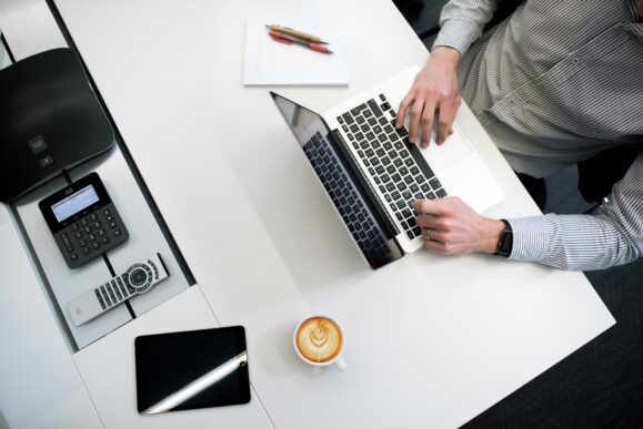 A business person working on a laptop with other devices and a cup of coffee shown on the desk | Phocas Software