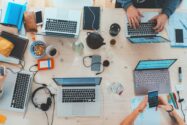 People sitting down near beige table with assorted laptop computers, headphones, coffee mugs, phones.