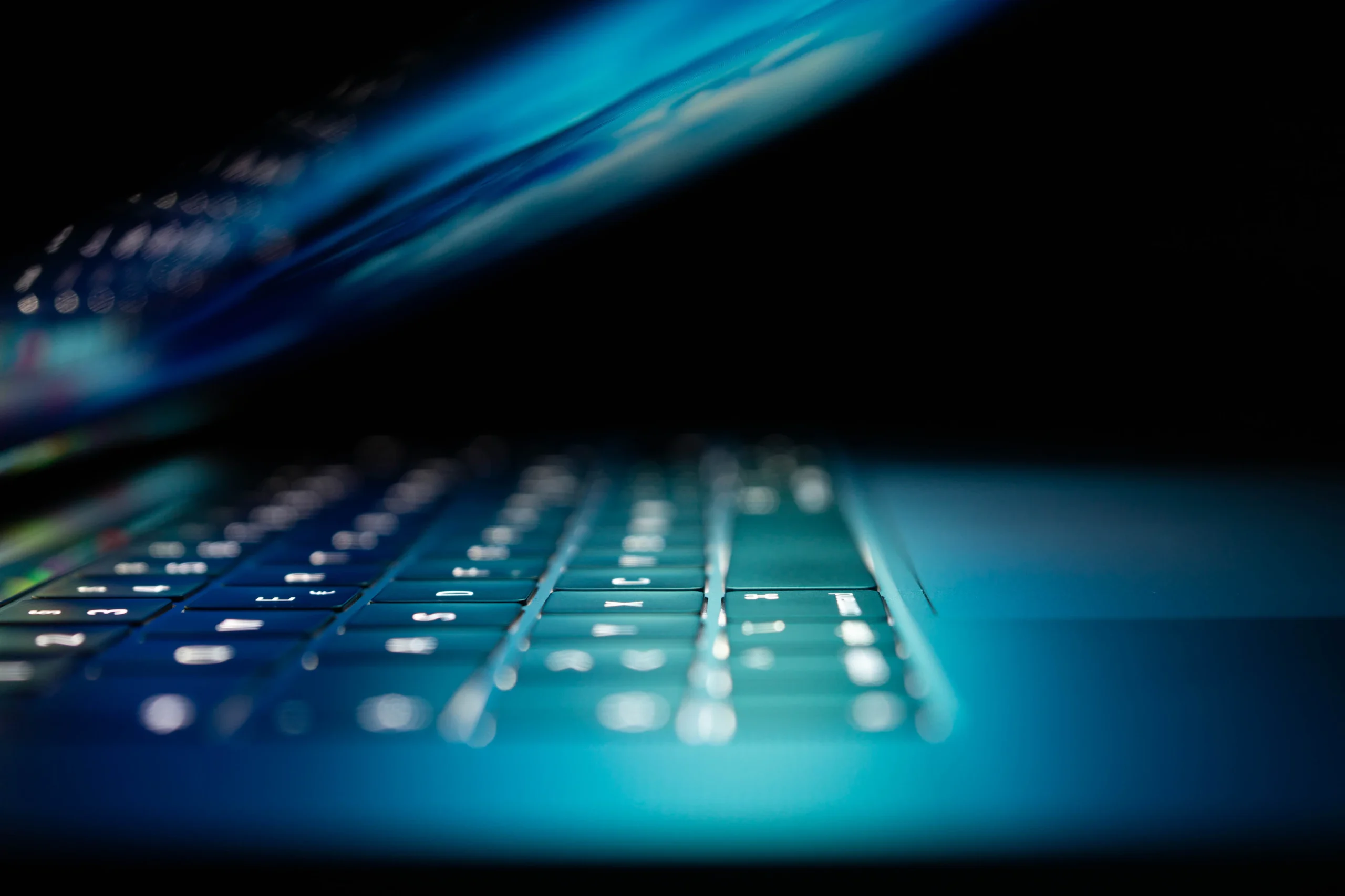 A lit up Mac laptop with keyboard shown in the close up. Blue light shines from the screen onto the keyboard.