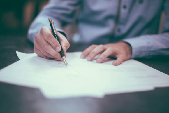 photograph of a man in a blue shirt signing a paper agreement/contract with a pen