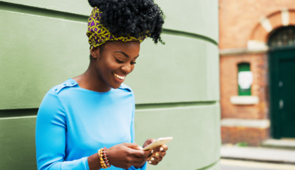 A happy female person with curly hair typing on her mobile phone. He stands outside in front of a green-colored wall