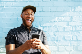 A happy male person wearing a cap and typing on his mobile phone. He stands in front of a light-blue wall