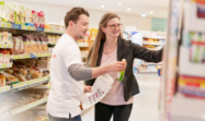 Two store employees talking with each other in a supermarket. The male employee on the left carries a printed staffing plan and a shelve product while the female colleague on the right is checking further on the shelve.