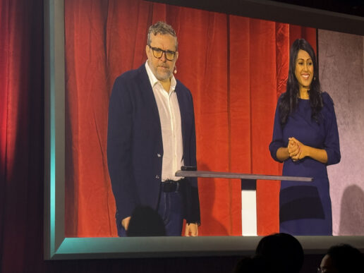 Two executives speaking on stage at an enterprise technology event, standing at a podium in front of a red curtain backdrop.