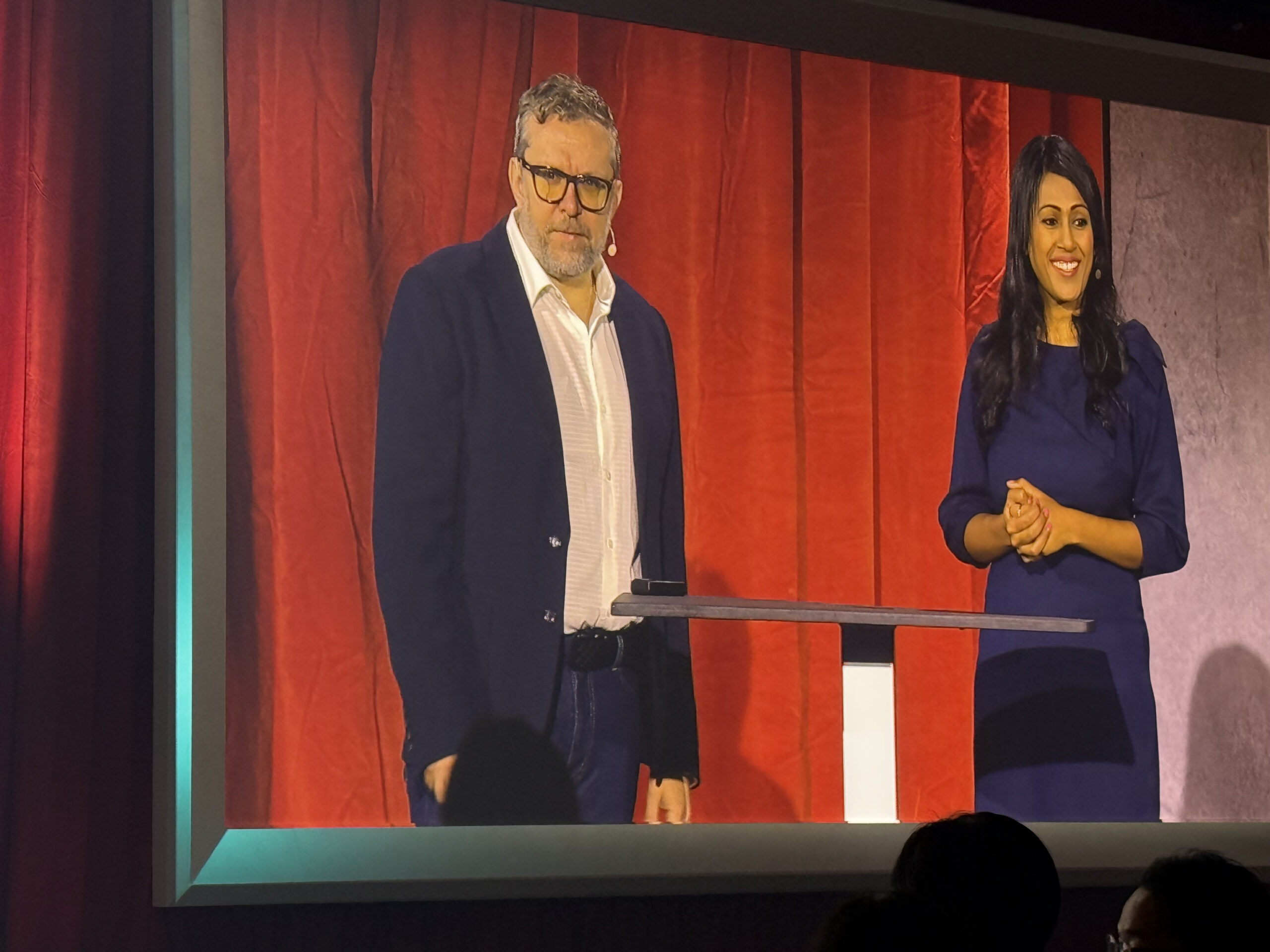 Two executives speaking on stage at an enterprise technology event, standing at a podium in front of a red curtain backdrop.