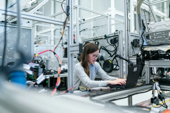 A female engineer in glasses working on a laptop at a wiring and electronics test station in a high-tech industrial facility, representing hands-on deployment of SAP Business AI, SAP Document AI, and BTP automation into live production systems.