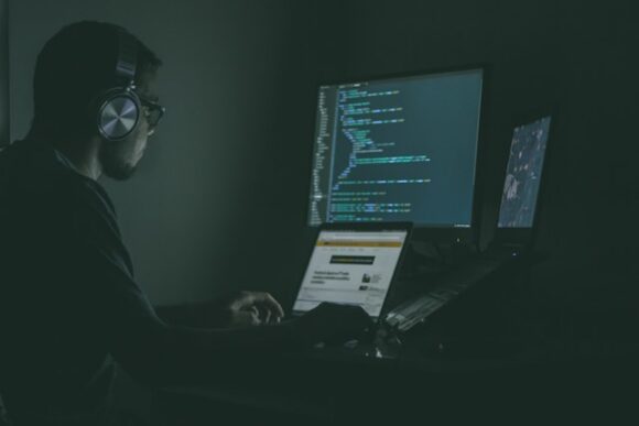 IT operations engineer wearing headphones monitoring code and system dashboards on multiple screens in a dark operations room, representing continuous SAP HANA high availability monitoring.
