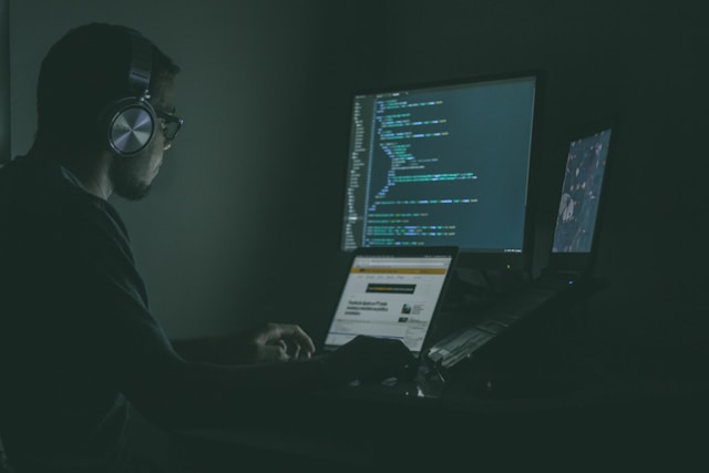 IT operations engineer wearing headphones monitoring code and system dashboards on multiple screens in a dark operations room, representing continuous SAP HANA high availability monitoring.