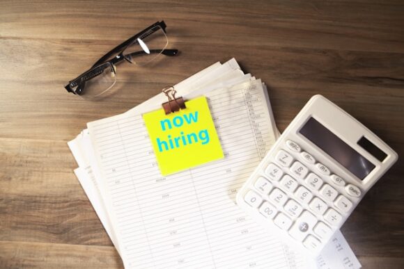 A bright yellow "now hiring" sticky note clipped to a stack of applicant tracking spreadsheets, beside reading glasses and a calculator on a wooden desk, representing the cost and volume of top-of-funnel recruiting that Radancy and SAP SuccessFactors aim to streamline with agentic AI.
