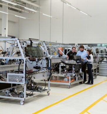 Three manufacturing professionals reviewing production data on a laptop and tablet beside modular assembly workstations on a clean, modern shop floor, representing SAP Digital Manufacturing connecting shop-floor execution to ERP-level planning on SAP BTP.