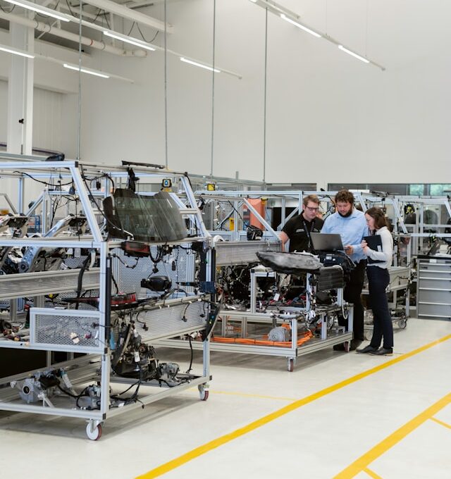 Three manufacturing professionals reviewing production data on a laptop and tablet beside modular assembly workstations on a clean, modern shop floor, representing SAP Digital Manufacturing connecting shop-floor execution to ERP-level planning on SAP BTP.