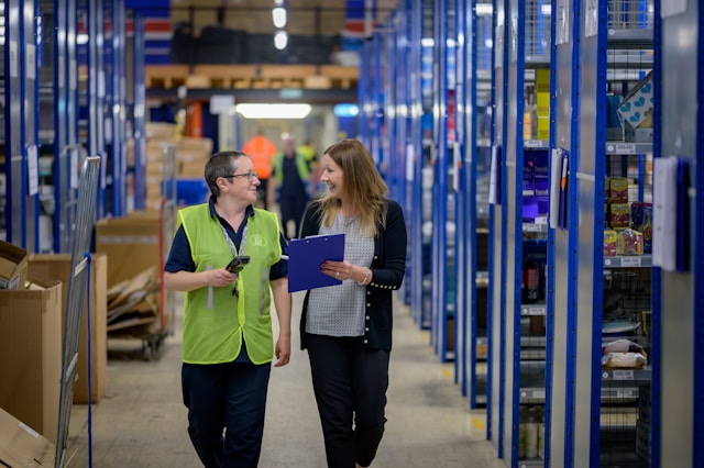 wo warehouse workers, one in a high-visibility vest holding a barcode scanner and one carrying a clipboard, walk between racking aisles, illustrating the SAP warehouse execution gap between the system of record and frontline work.