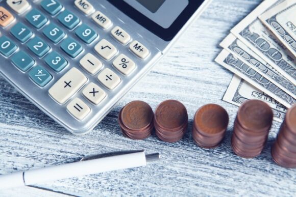 Calculator, stacked coins, banknotes, and a pen on a desk, representing the manual pay calculations that SAP SuccessFactors Payroll replaces with continuous, automated payroll processing.