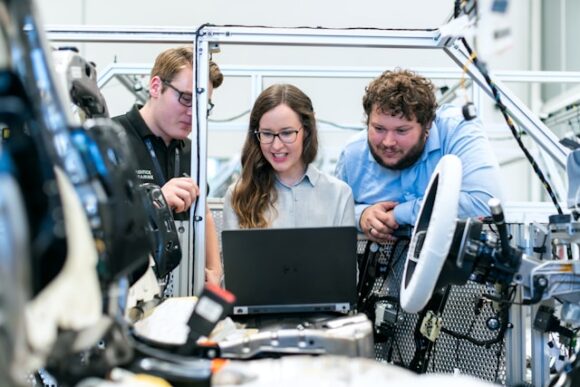 Three automotive engineers review sourcing and engineering data on a laptop beside a vehicle prototype chassis, illustrating the digital thread connecting product design to SAP procurement execution.