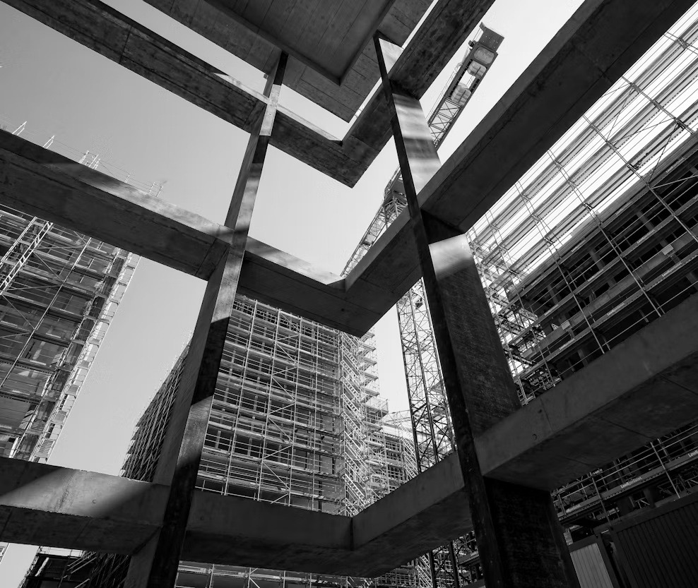Black-and-white view of a building under construction, with exposed concrete beams, scaffolding, and cranes.