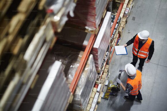 Warehouse workers review inventory beside stacked pallets, illustrating the operational complexity behind pricing, rebates, and margin visibility.